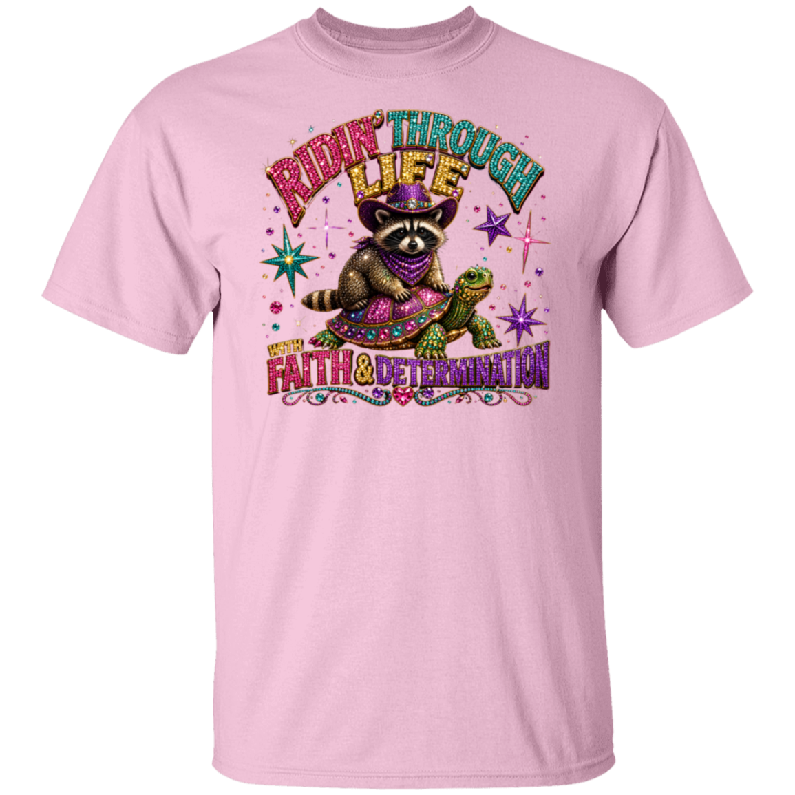 A light pink short-sleeve crewneck t-shirt featuring a colorful graphic of a raccoon wearing a purple hat and scarf, riding a green turtle. The graphic includes the text 'RIDE THROUGH LIFE' and 'WONDER FAITH & DETERMINATION' in stylized fonts with decorative sparkles.