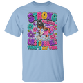 A light blue short-sleeve crewneck t-shirt featuring a colorful graphic print on the front. The graphic displays the text 'STRONG FOCUSED UNSTOPPABLE THAT'S MY VIBE' in a playful, multi-colored bubble font, accompanied by an illustration of a woman with braided hair in a pink dress and a small white dog surrounded by flowers.