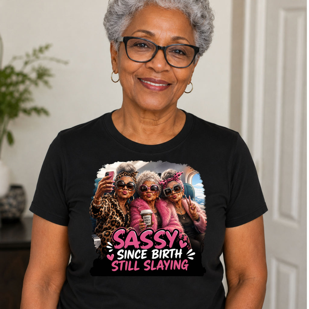 An older woman with short gray hair and glasses wearing a black graphic t-shirt. The shirt features a colorful print of three women taking a selfie with the text 'SASSY SINCE BIRTH STILL SLAYING' in pink and white.