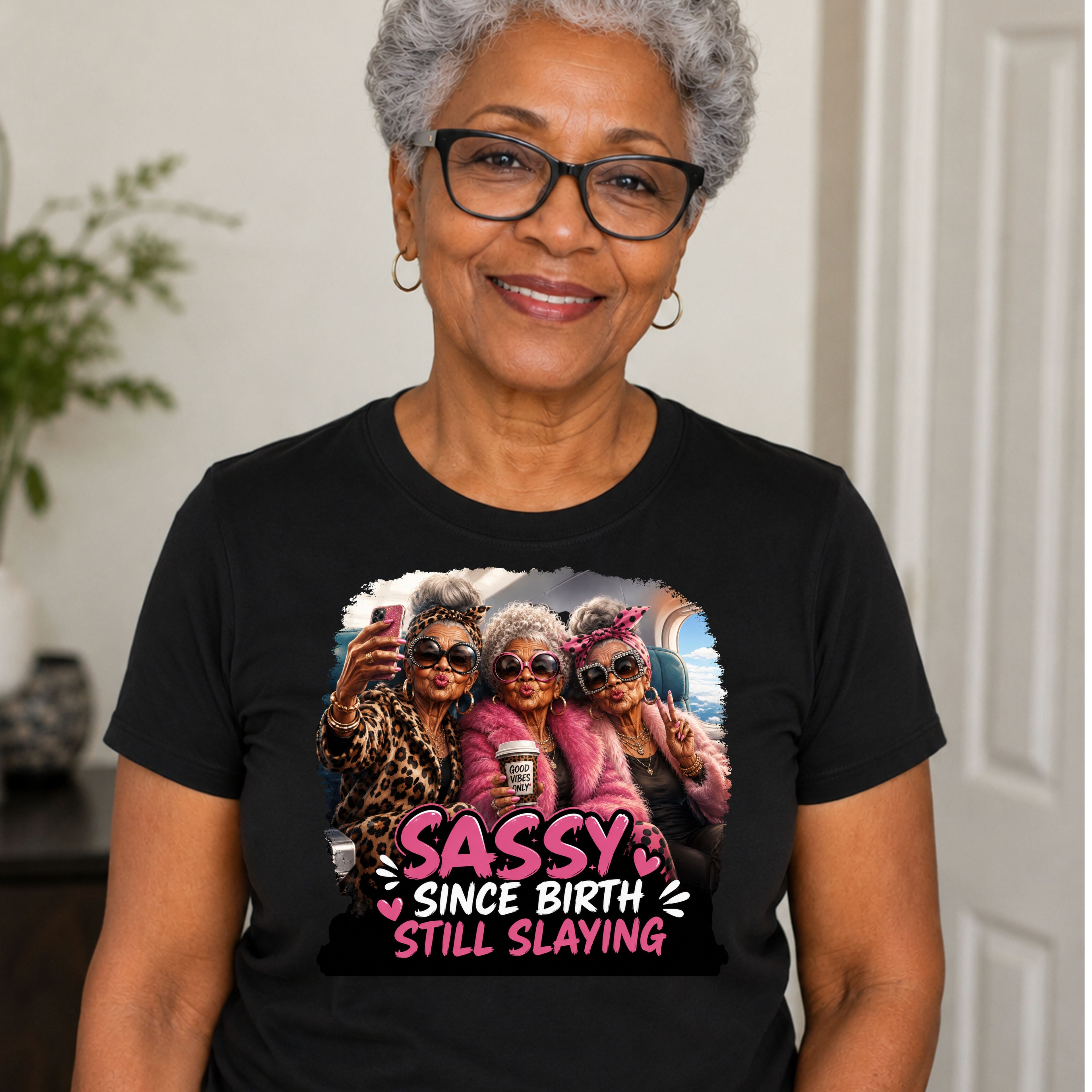 An older woman with short gray hair and glasses wearing a black graphic t-shirt. The shirt features a colorful print of three women taking a selfie with the text 'SASSY SINCE BIRTH STILL SLAYING' in pink and white.
