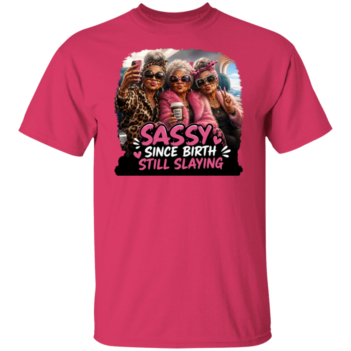 A heliconia color short-sleeve crewneck t-shirt featuring a large graphic print on the front. The graphic shows three elderly women taking a selfie with sunglasses and the text 'SASSY SINCE BIRTH STILL SLAYING' in stylized pink and white lettering.