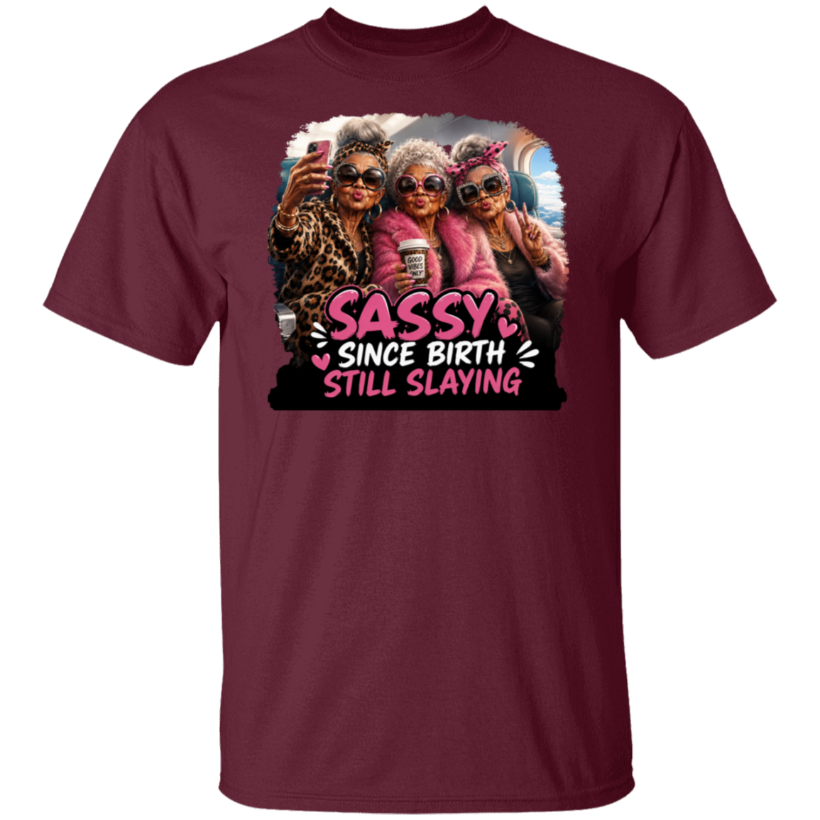 A maroon short-sleeve crewneck t-shirt featuring a colorful graphic print of three women taking a selfie with the text 'SASSY SINCE BIRTH STILL SLAYING' in pink and white.