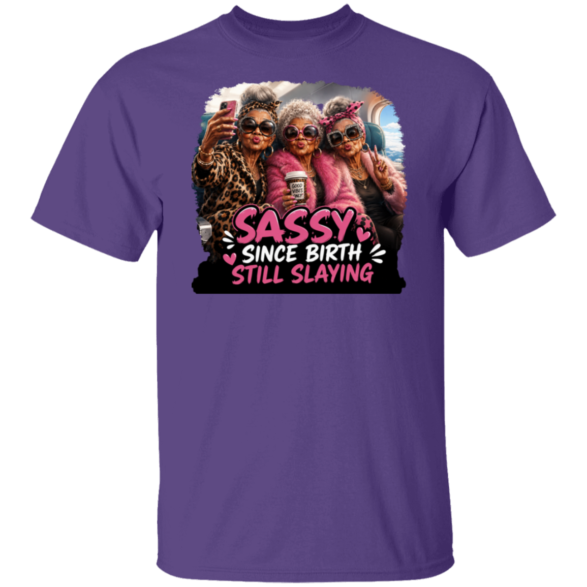 A purple short-sleeve crewneck t-shirt featuring a large graphic print on the front of three elderly women taking a selfie with the text 'SASSY SINCE BIRTH STILL SLAYING' in pink and white.