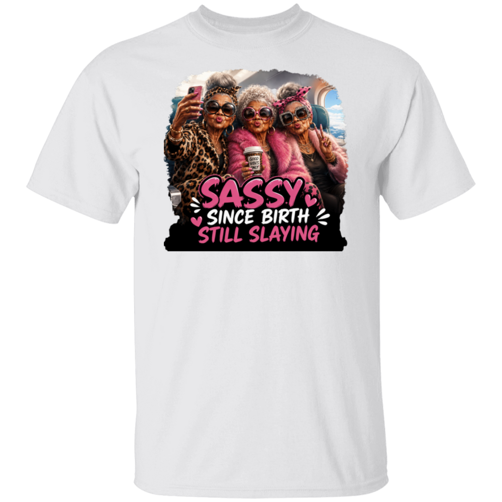 A white short-sleeve crewneck t-shirt featuring a colorful graphic print of three elderly women taking a selfie with the text 'SASSY SINCE BIRTH STILL SLAYING' in pink and white lettering.