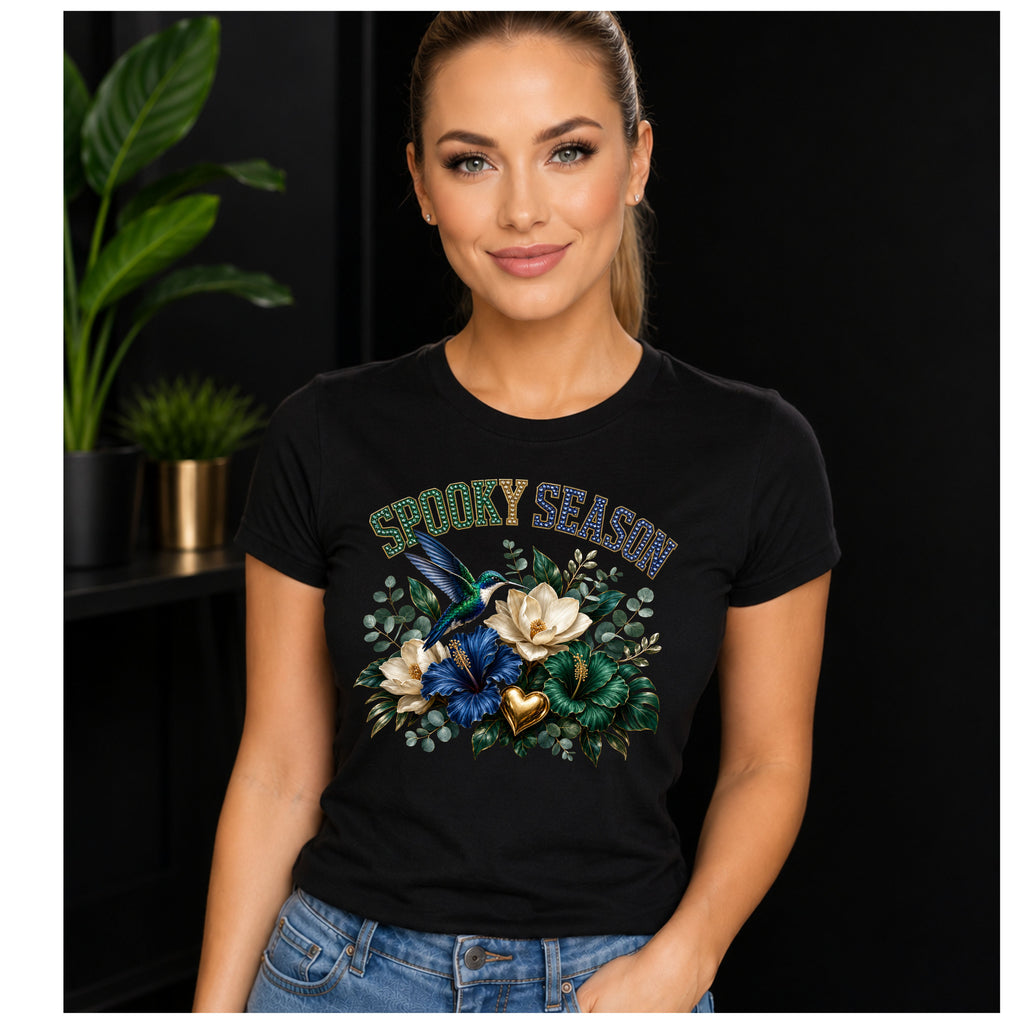 A woman wearing a black graphic t-shirt with a 'Spooky Season' design featuring a blue bird, white flowers, and green leaves, set against a dark background with indoor plants.