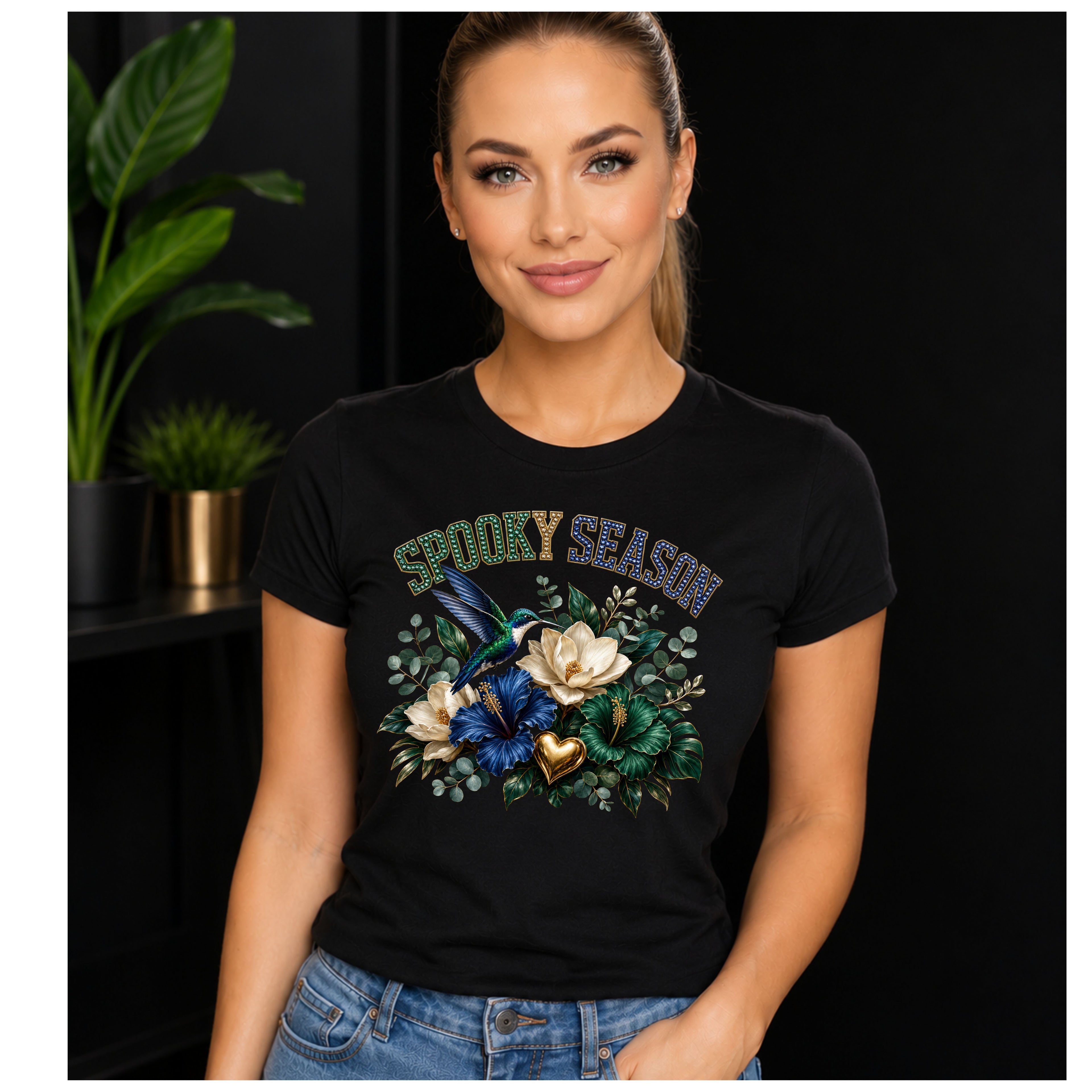 A woman wearing a black graphic t-shirt with a 'Spooky Season' design featuring a blue bird, white flowers, and green leaves, set against a dark background with indoor plants.