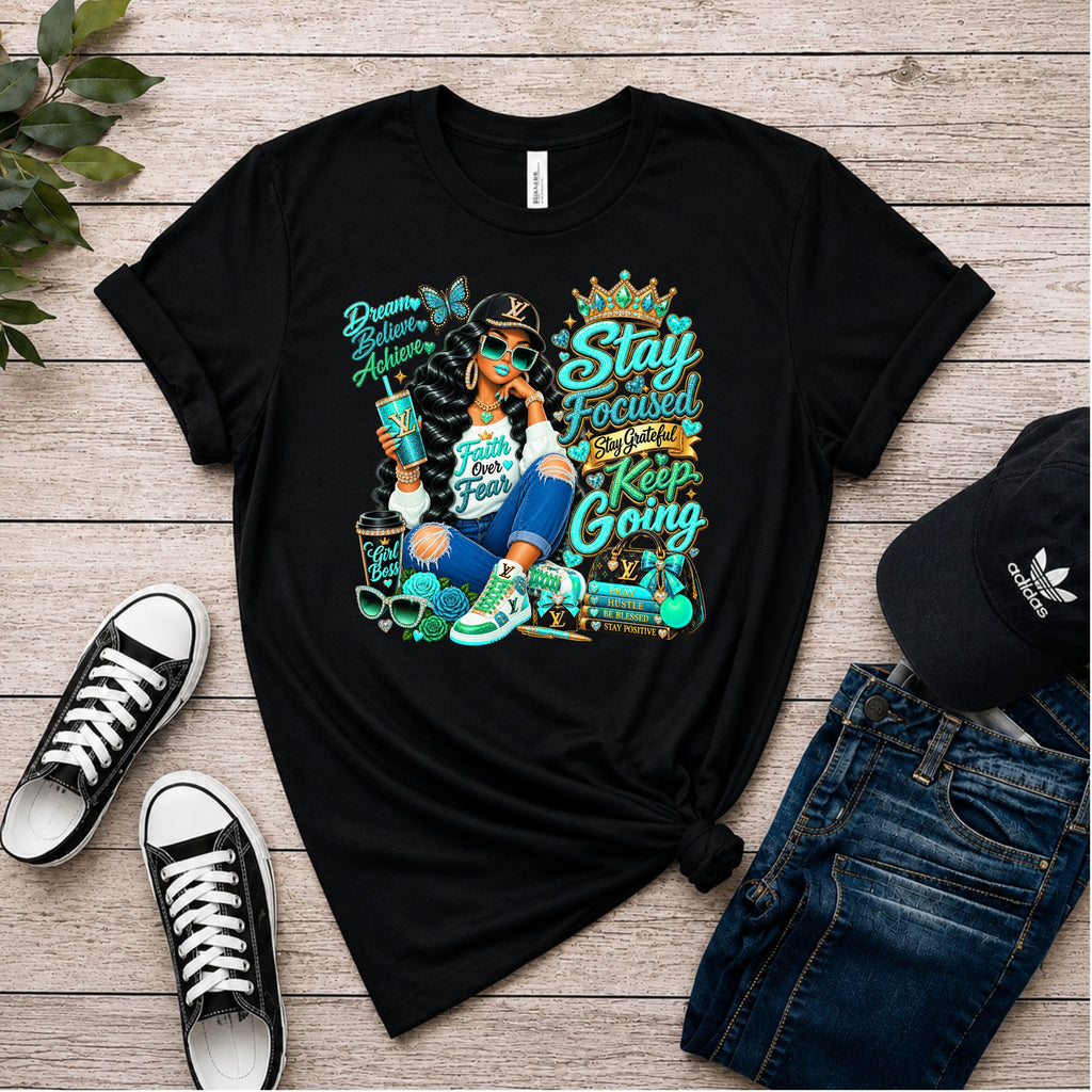 A black graphic t-shirt laid flat on a light wood plank background, styled with black sneakers, blue jeans, and a black Adidas cap. The t-shirt features a colorful graphic of a woman with a crown and motivational text including 'Stay Focused', 'Keep Going', and 'Dreams Become Reality'.