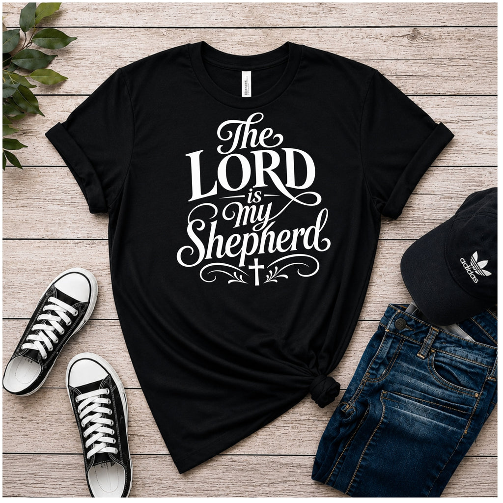 A black color graphic T-shirt laid flat on a rustic wood plank surface, styled with black sneakers, blue jeans, and a black Adidas cap. The shirt has a large white typographic design reading “The LORD is my Shepherd” with decorative flourishes and a small cross.