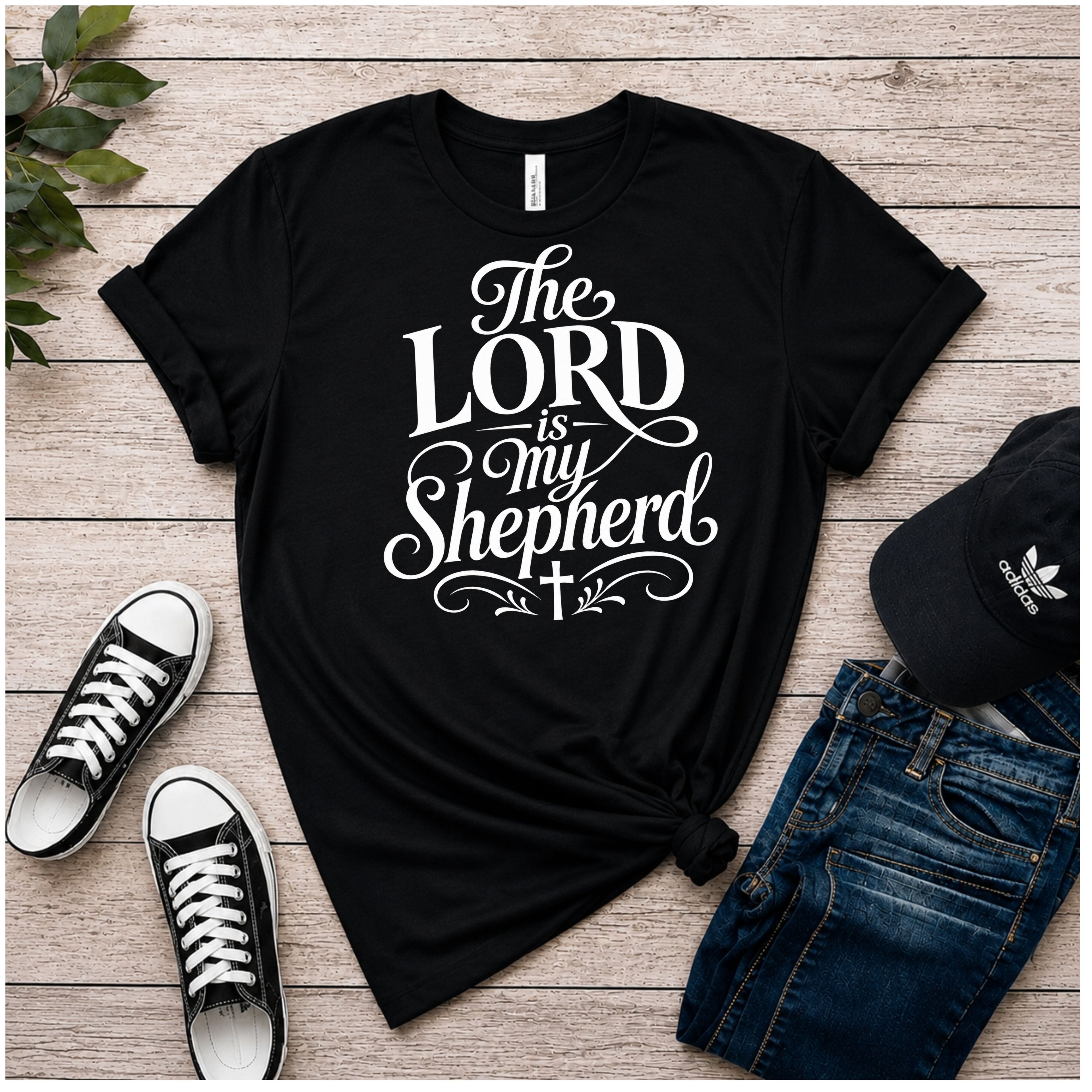 A black color graphic T-shirt laid flat on a rustic wood plank surface, styled with black sneakers, blue jeans, and a black Adidas cap. The shirt has a large white typographic design reading “The LORD is my Shepherd” with decorative flourishes and a small cross.