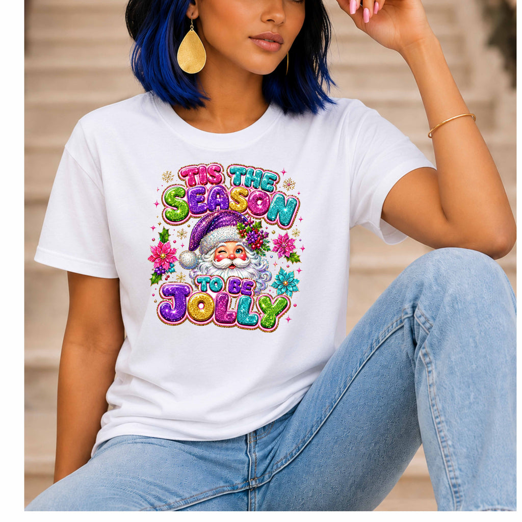 A woman wearing a white short-sleeve graphic T-shirt with a colorful festive Santa graphic and the text 'IT'S THE SEASON TO BE JOLLY', paired with light blue jeans, seated against a blurred beige background.