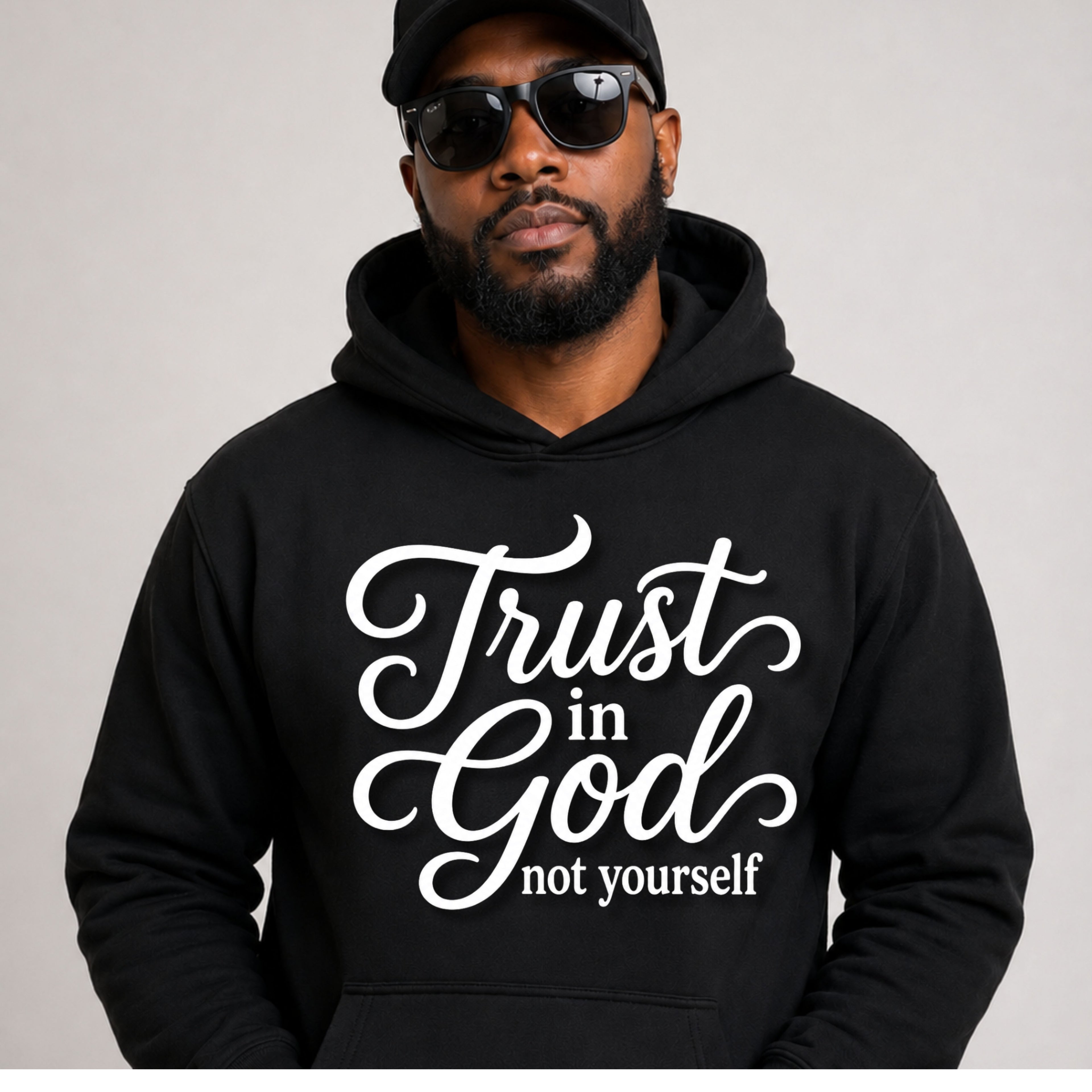 A man wearing a black hoodie with the white text 'Trust in God not yourself' on the front, along with a black cap and sunglasses, against a plain light gray background.