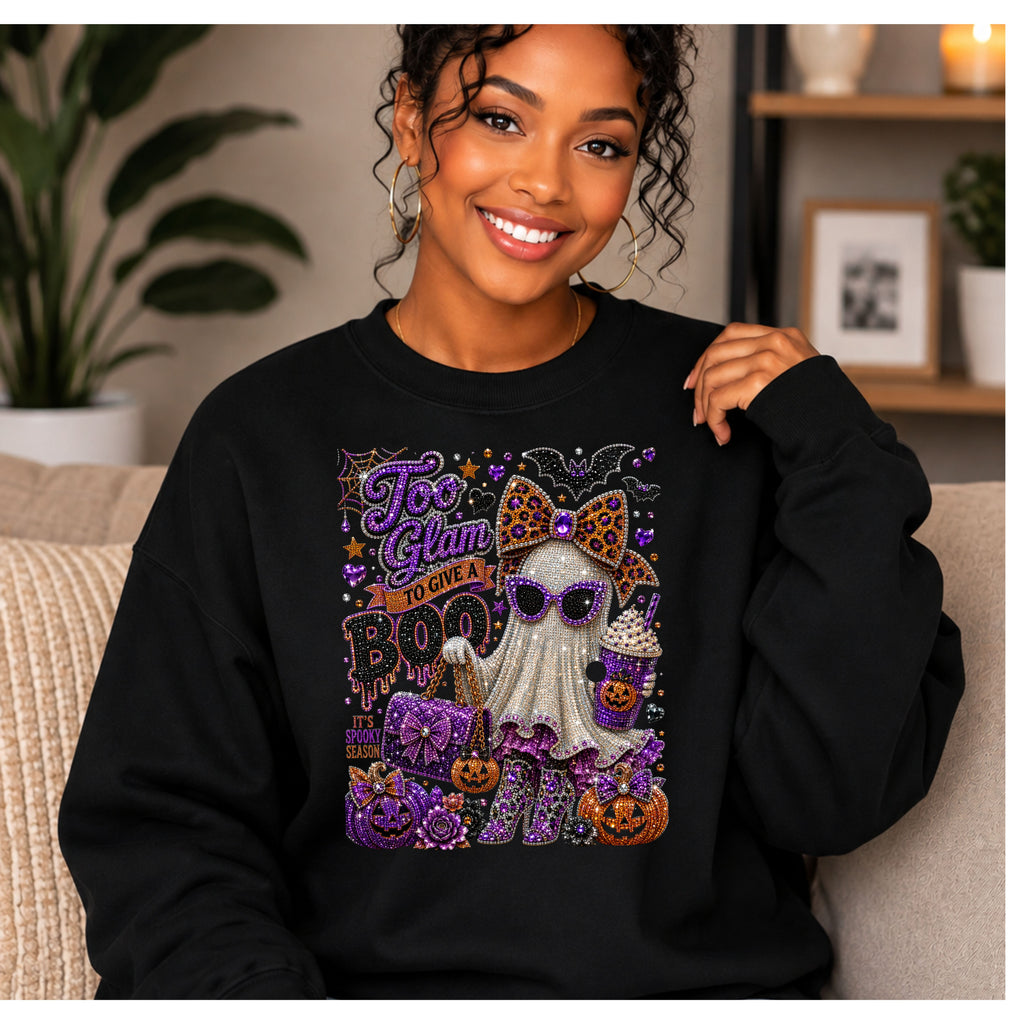 A woman wearing a black crewneck sweatshirt with a large, glittery Halloween graphic on the front. The graphic features a ghost character with a leopard-print bow, surrounded by pumpkins, a bat, and the text 'Too Glam to Care' and 'BOO'.
