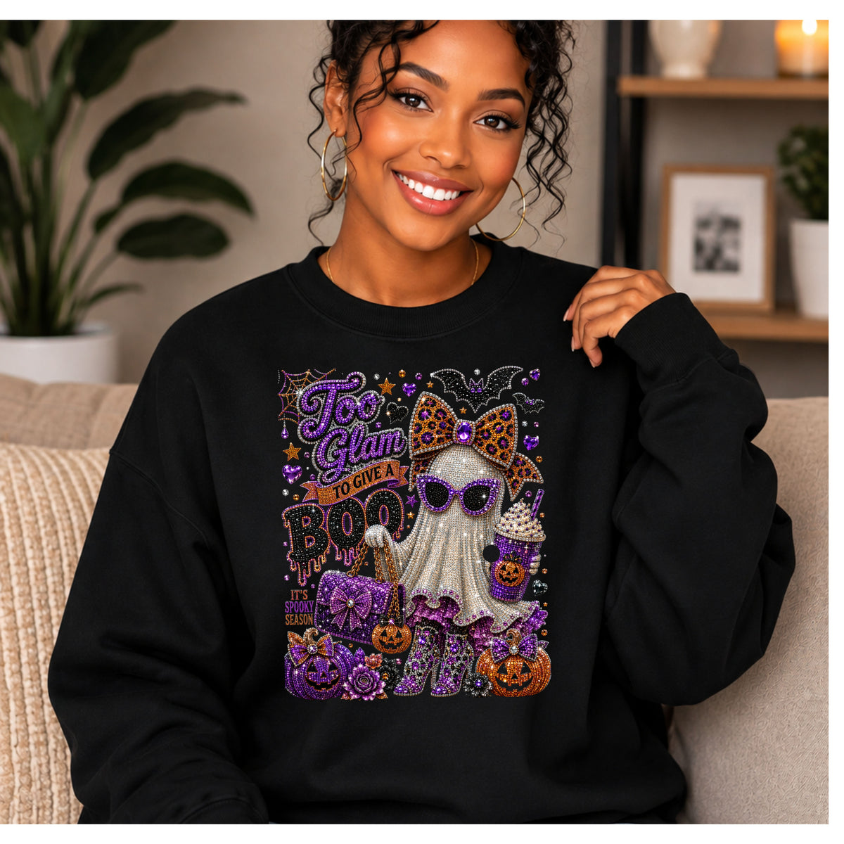 A woman wearing a black crewneck sweatshirt with a large, glittery Halloween graphic on the front. The graphic features a ghost character with a leopard-print bow, surrounded by pumpkins, a bat, and the text 'Too Glam to Care' and 'BOO'.