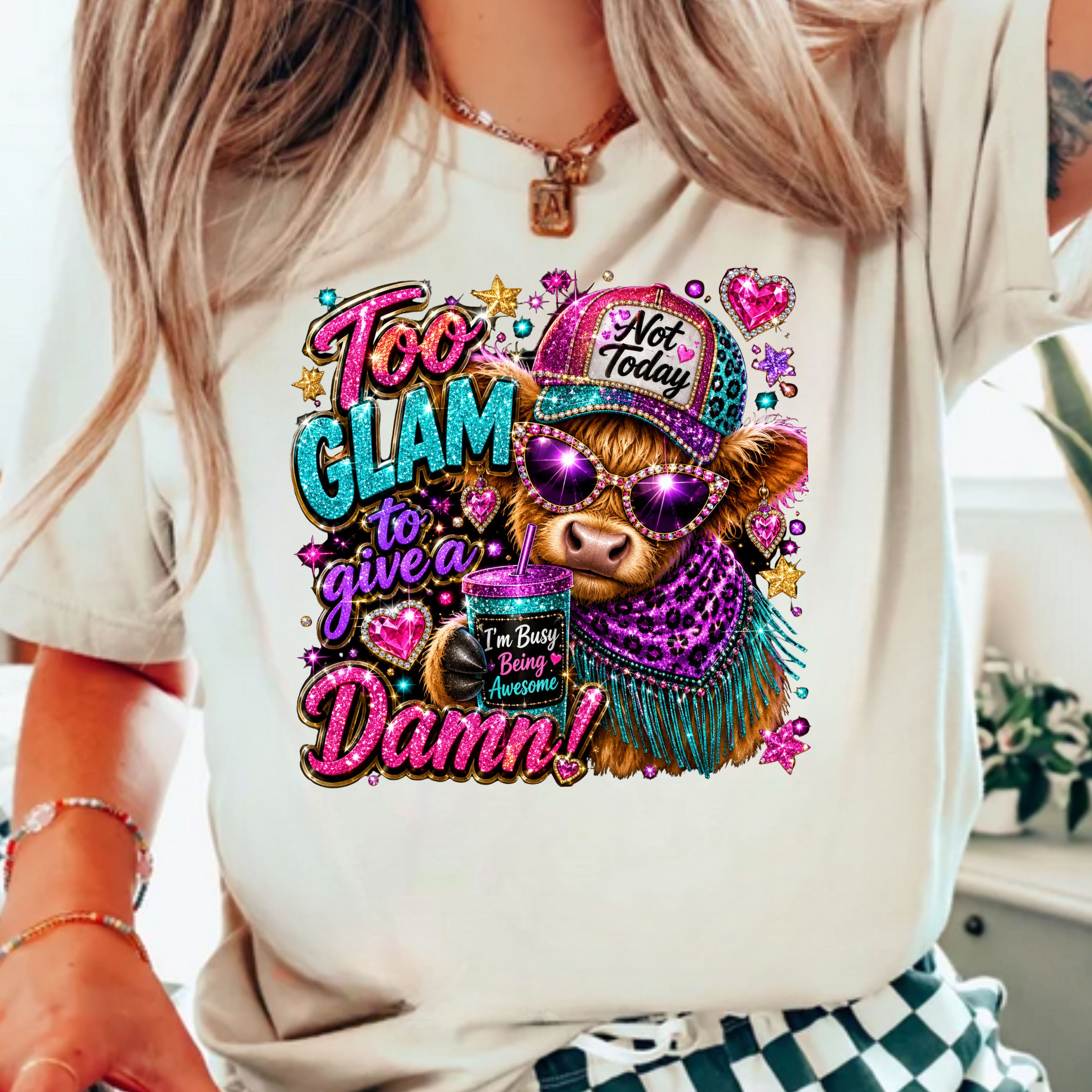 A woman wearing a cream-colored graphic t-shirt with a colorful design of a cartoon cow wearing a purple hat and scarf, surrounded by text that reads 'Too GLAM to give a Damn!'.