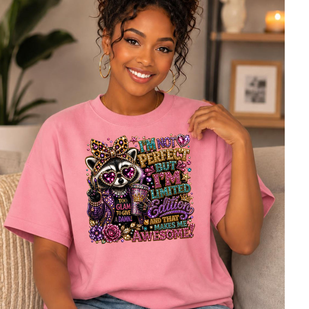 A smiling woman wearing a pink graphic T-shirt with a colorful raccoon and glitter-style text design, seated indoors on a light-colored couch with a plant and framed decor in the background.