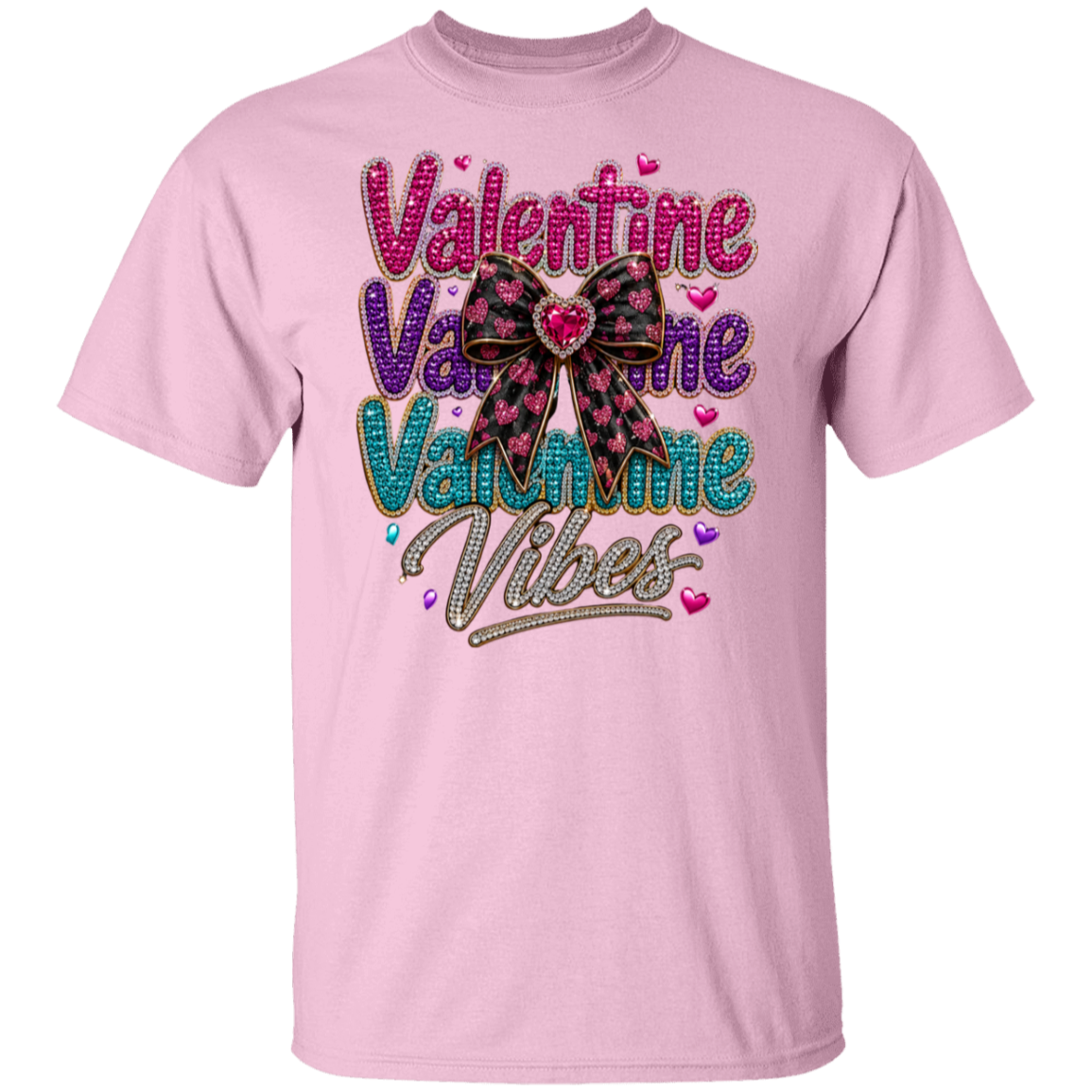 A light pink short-sleeve crewneck t-shirt featuring a colorful graphic on the front with the text 'Valentine Valentine Valentine Vibes' in pink, purple, teal, and silver glitter-style lettering, accented by a pink and black plaid bow with a red heart in the center.