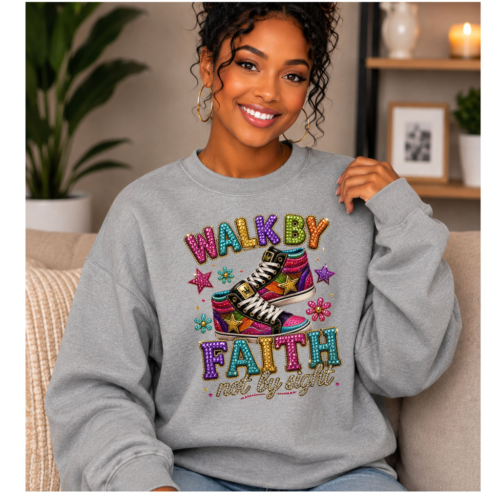 A woman wearing a light gray crewneck sweatshirt with a colorful graphic that reads 'WALK BY FAITH not by sight' above and below a pair of high-top sneakers. She is sitting on a beige couch in a cozy living room setting.