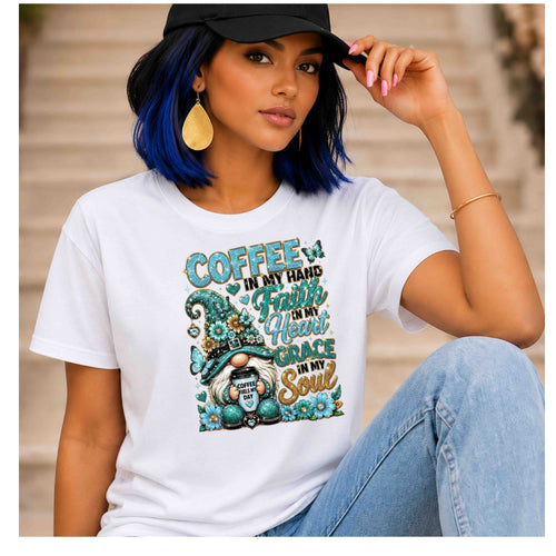 A woman wearing a white graphic t-shirt with a teal and gold 'Coffee in my hand, Faith in my heart, Grace in my soul' gnome design, paired with a black cap and light blue jeans.