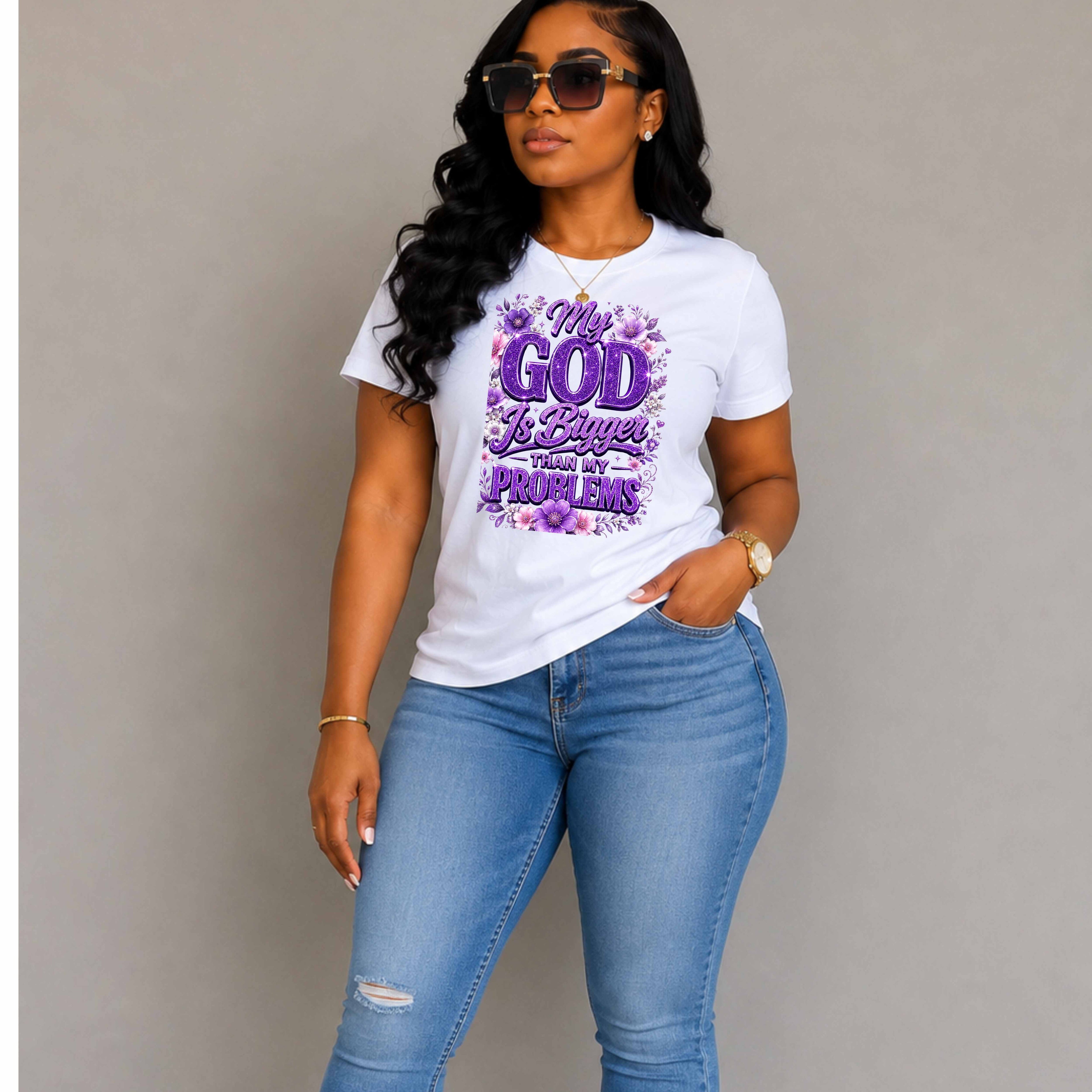 A woman modeling a white graphic t-shirt with purple text that reads 'My GOD Is Greater Than My Problems' surrounded by purple flowers, paired with light blue jeans against a plain gray background.