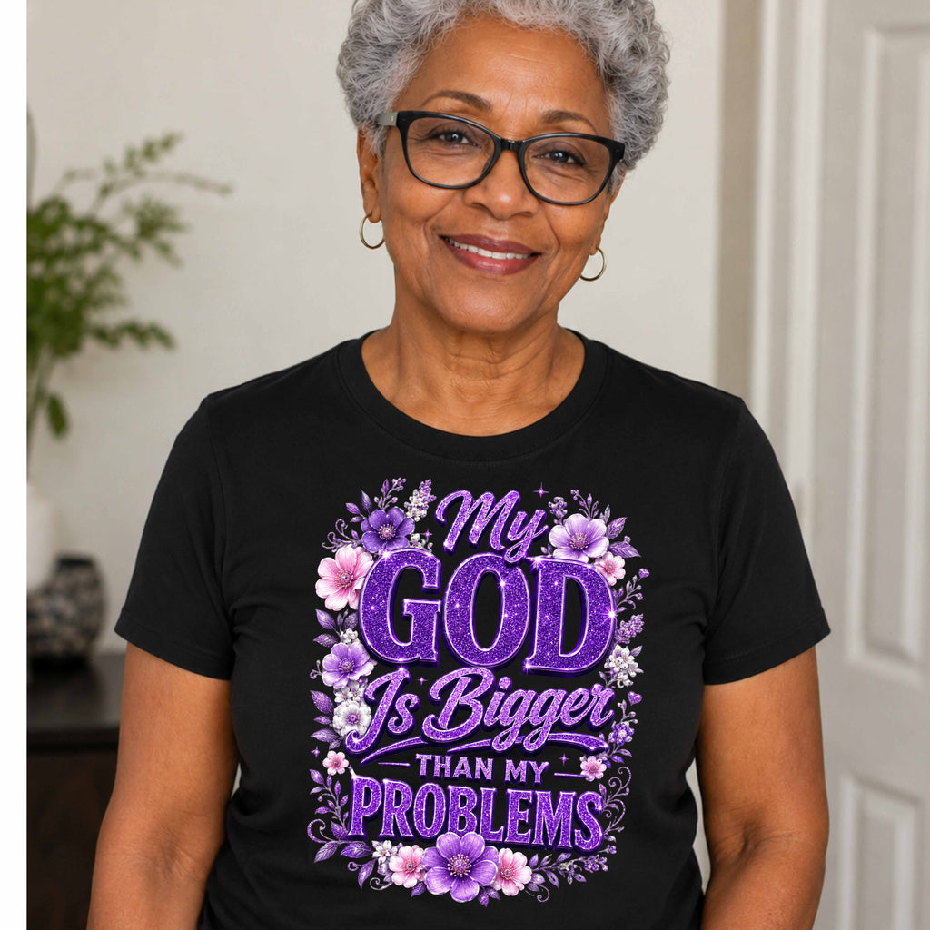 An older woman with short gray hair and glasses is shown from the chest up, wearing a black graphic T-shirt with purple floral artwork and the text 'My GOD Is Bigger THAN MY PROBLEMS' on the front.
