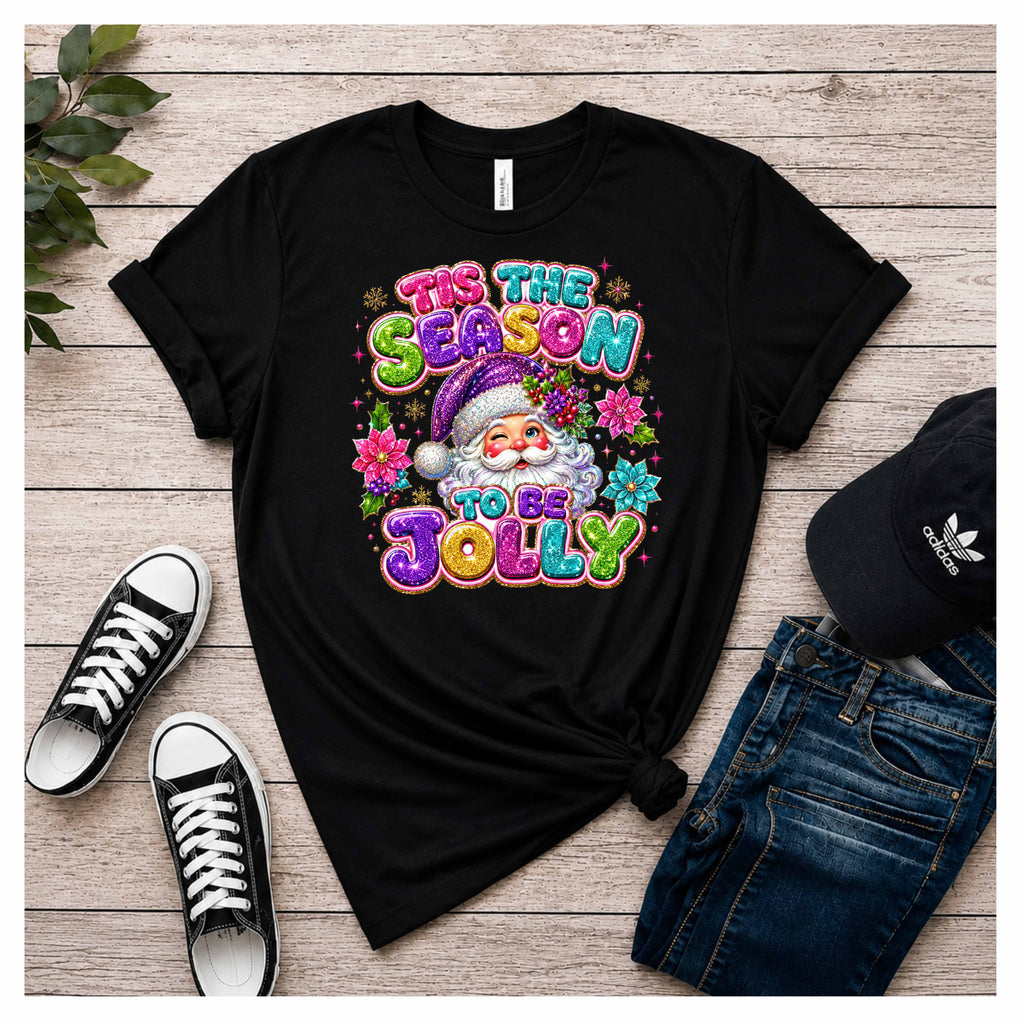 A black color graphic t-shirt featuring a colorful Christmas design with a Santa Claus illustration and the text 'TIS THE SEASON TO BE JOLLY' in pink, green, purple, and yellow. The t-shirt is styled in a flat lay on a light wood background, accompanied by black sneakers, blue jeans, and a black Adidas cap.